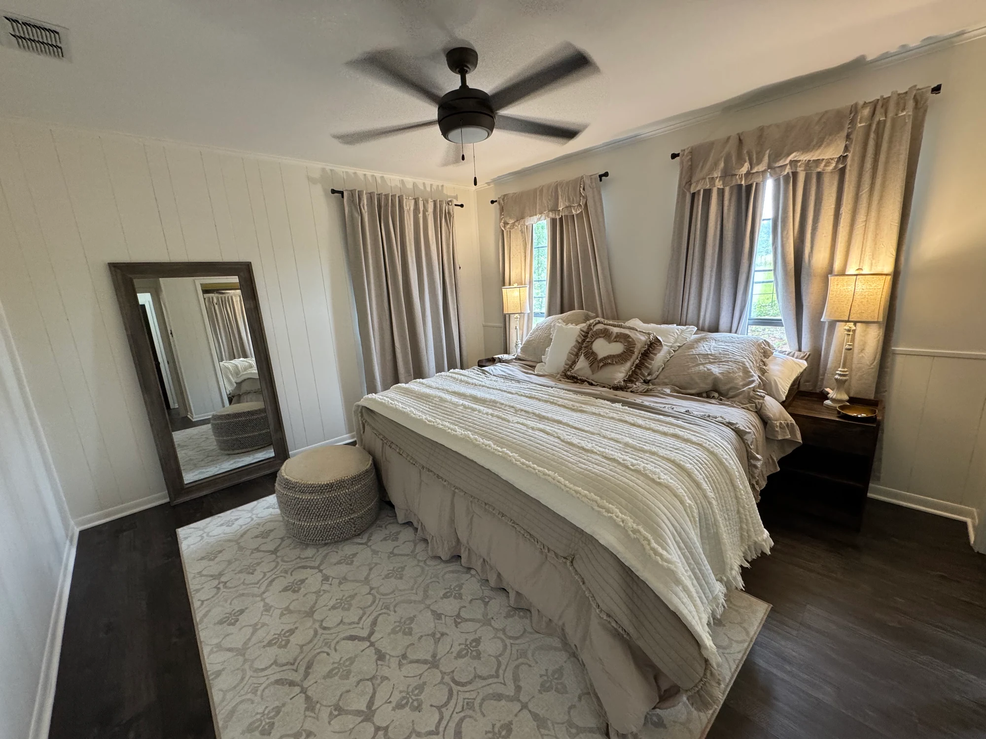 Bedroom with neutral bedding, a leaning mirror and curtained windows